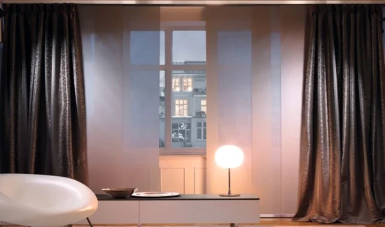 Dunkelbraune blickdichte Flächenvorhänge vor einem abendlichen Fenster in einem schwach beleuchteten Raum