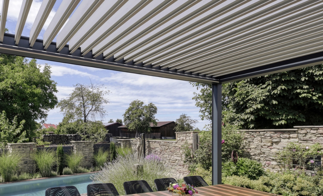 Sitzgruppe unter einem Lamellendach auf einer Terrasse neben einem Pool. Der Garten wird von einer halbhohen Mauer umzäunt.